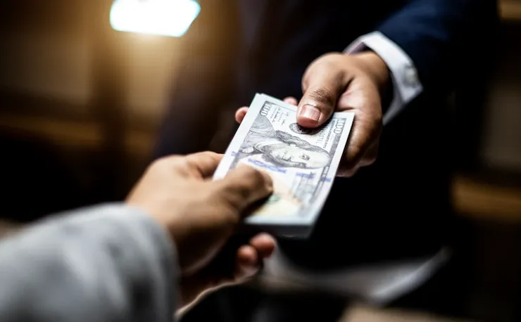 Fotografía en primer plano de la mano de un empresario entregando un fajo de 100 billetes de dólar a otra persona