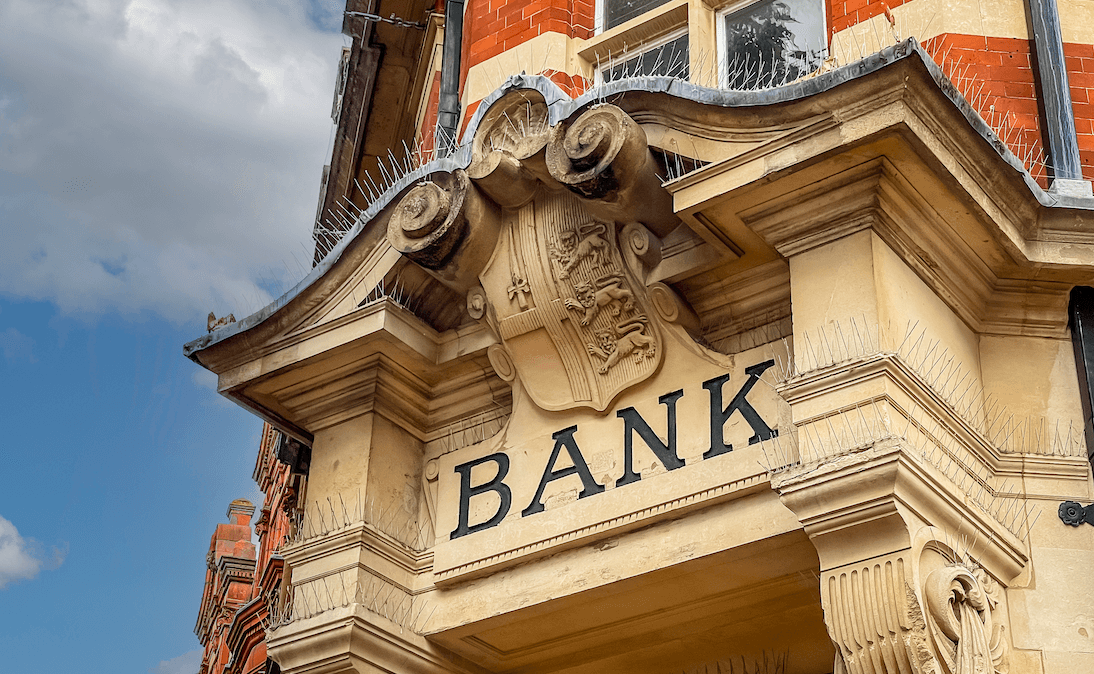 The stone facade of a British bank, with ornate scrollwork and the word "Bank" engraved over the doorway.
