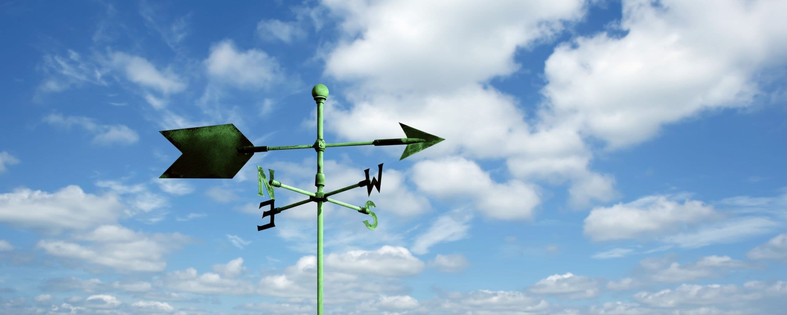 A weather vane stands against a bright blue sky with puffy white clouds.