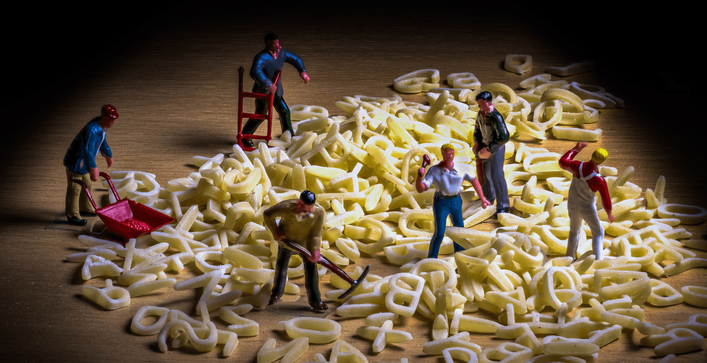 Una fotografía de figuritas en miniatura que representan a unos obreros con picos y carretillas excavando en un montículo de letras que forman una «sopa de letras».