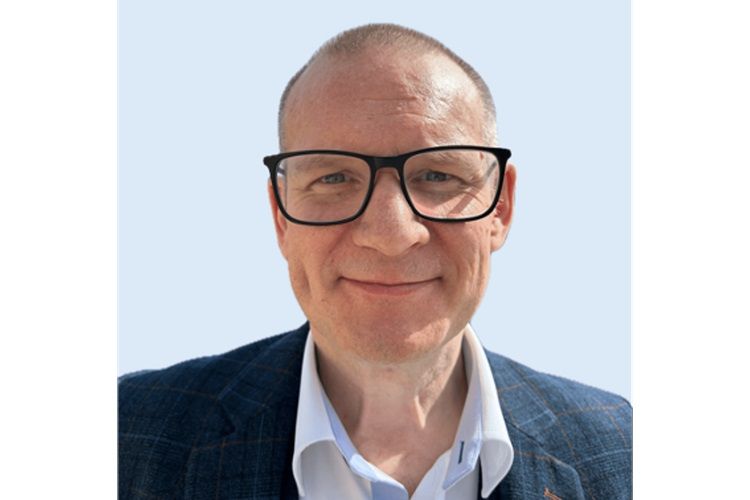 Imagen de un hombre blanco con el pelo muy corto y gafas de montura grande, que lleva una chaqueta gris a cuadros.