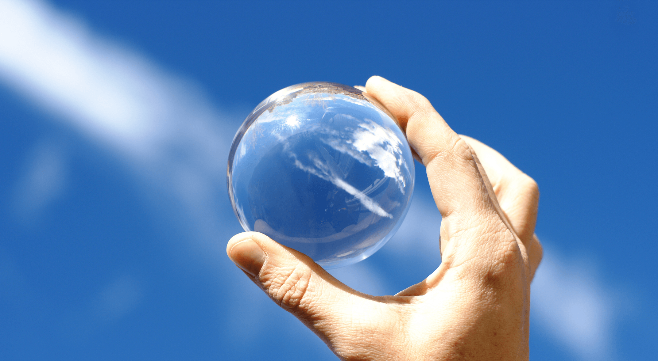 Una mano sostiene una bola de cristal en alto contra un cielo azul con nubes tenues.