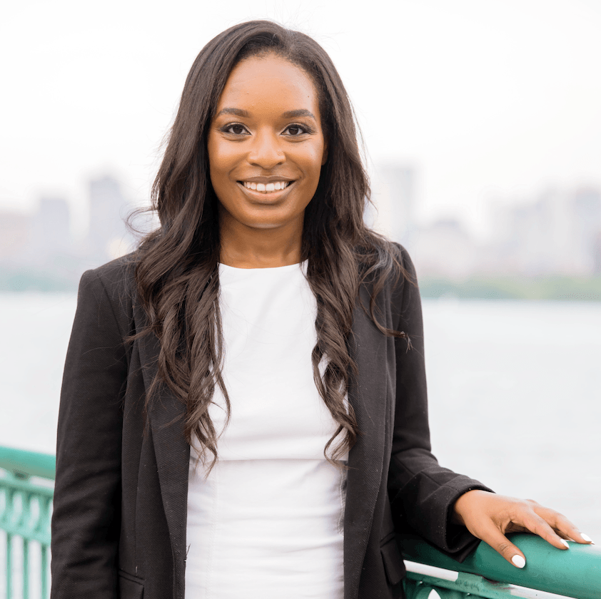 Fotografía de una mujer negra con el pelo largo y liso, vestida con una camiseta blanca y una chaqueta negra, de pie junto a la barandilla de un canal urbano.