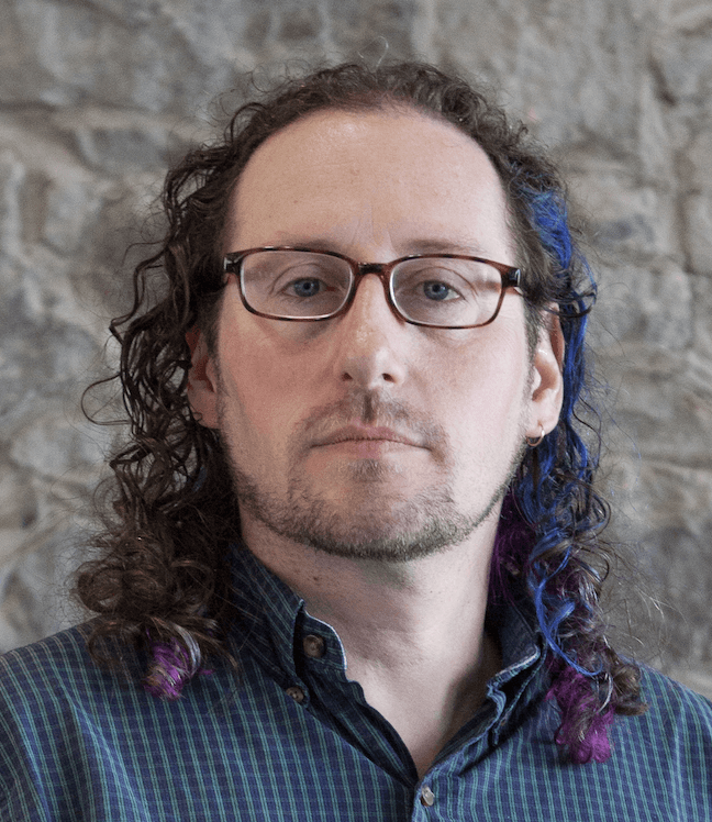 Fotografía del autor. Un hombre blanco de unos 30s o 40s años, con gafas y un mechón azul entre su pelo castaño y rizado.