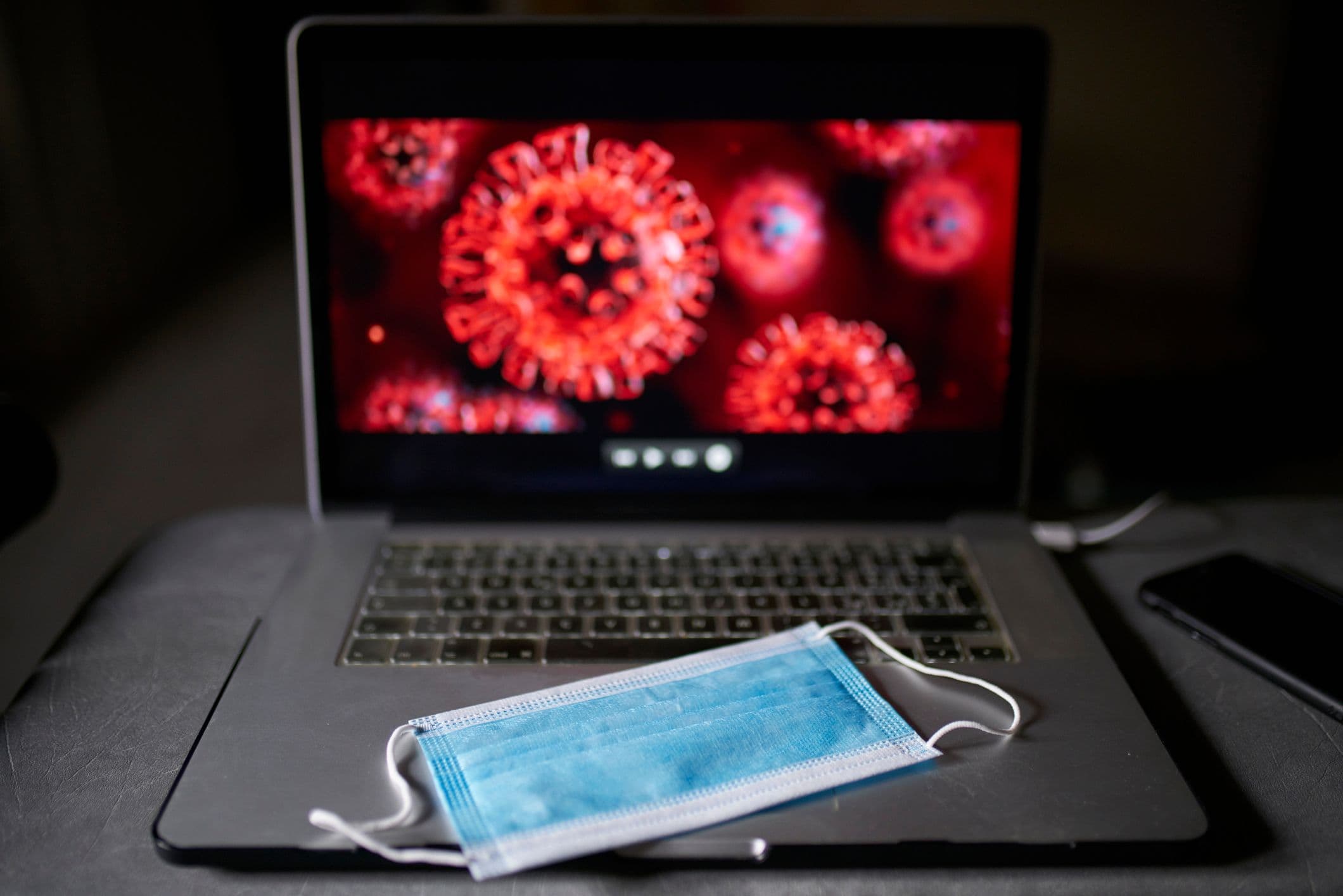 In a dark space, a blue face mask sits on a laptop keyboard, with microscopic images of the spiky red COVID virus glaring from the screen.