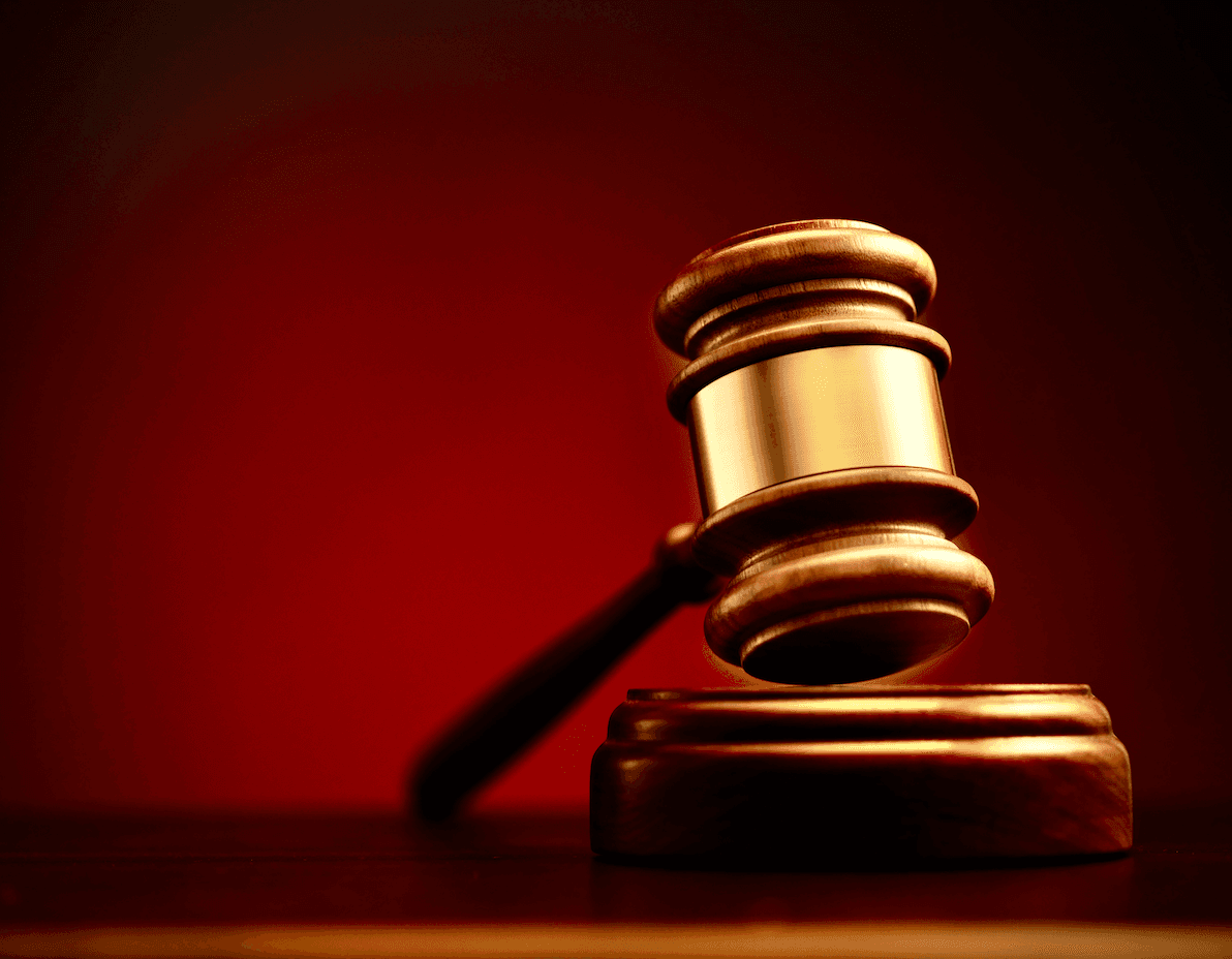 A polished wooden gavel with gold trim rests on a sound block against a crimson background.